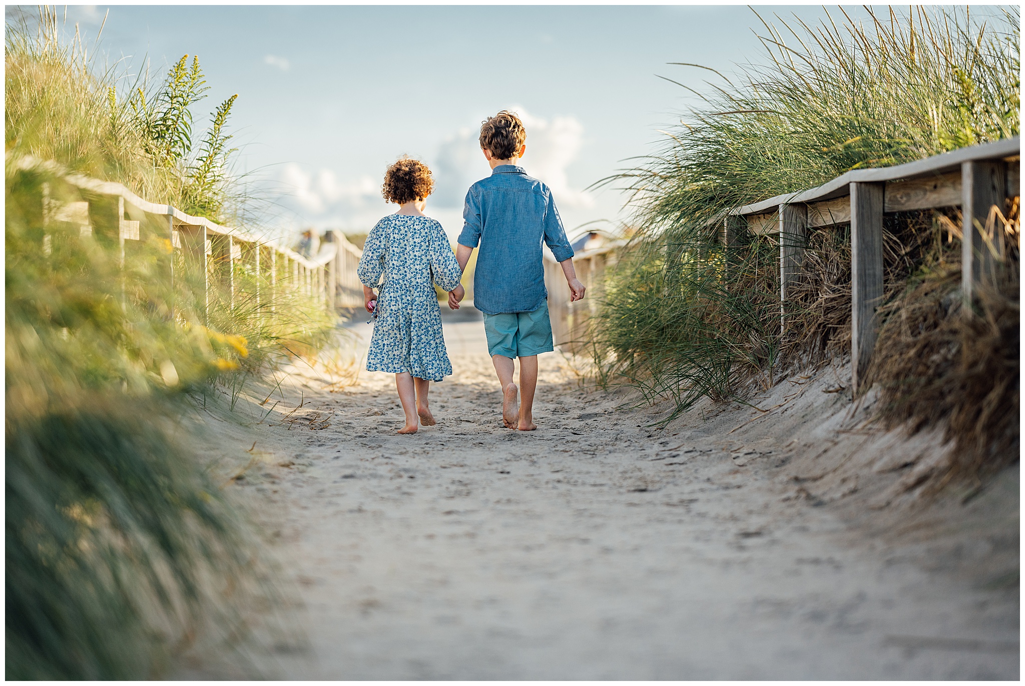 A photograph of two siblings (a little girl and a little boy) holding hands and walking away from the camera at the beach during a family photography and videography session in the Boston area.