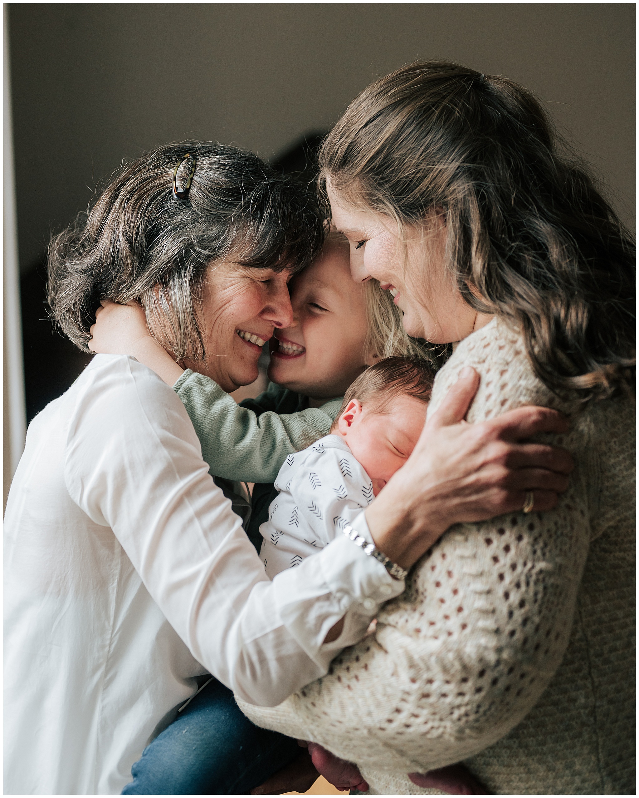 A professional photograph of a grandmother with her arms wrapped around her daughter and her two grandchildren. They are all smiling and happy.